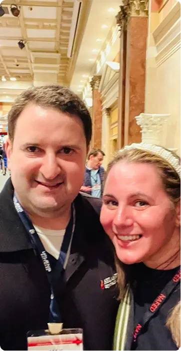 Smiling man and woman posing for a selfie at a conference, wearing lanyards and badges in a decorative hallway.
