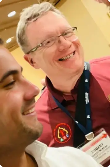 Two men smiling at an event; the man in maroon with a lanyard and badge showing the name Robert.