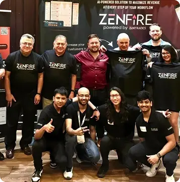Group of people in ZenFire shirts posing together at a conference or event, with a ZenFire banner and presentation screens in the background.