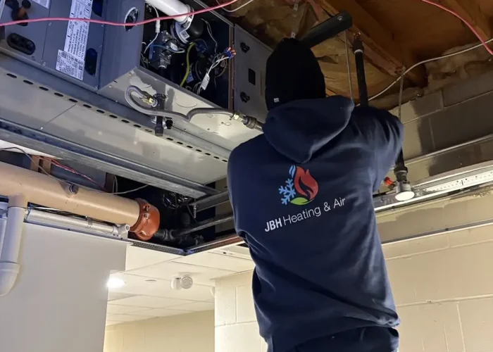 HVAC technician in a JBH Heating & Air hoodie stands on a ladder, working on an exposed furnace and wiring in a ceiling space.